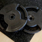 Two black weight plates on top of gray speckled rubber gym tiles.