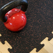 Red kettlebell with black handle on a terra cotta speckled rubber gym tile.
