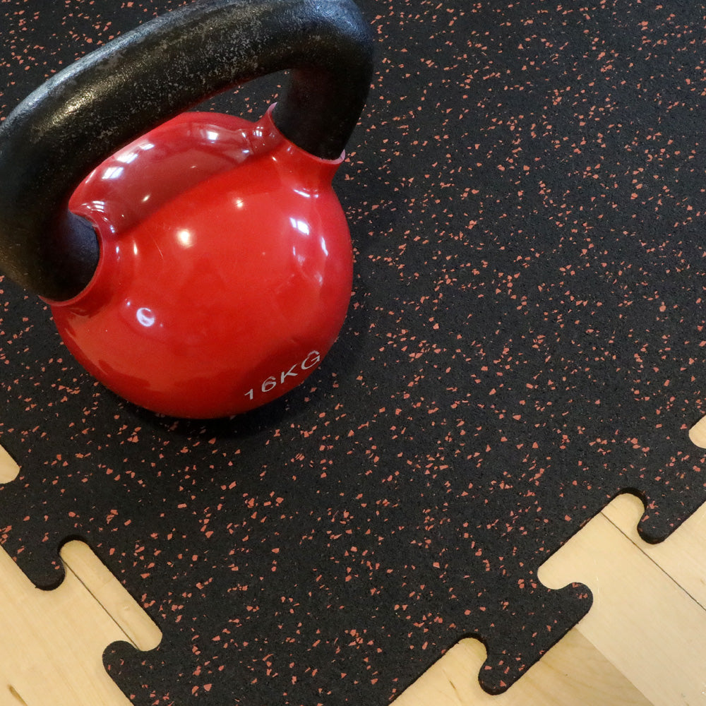 Red kettlebell with black handle on a terra cotta speckled rubber gym tile.
