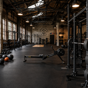 A spacious, industrial-style gym interior featuring professional fitness equipment including power racks with barbells and bumper plates, a rowing machine, and a variety of kettlebells and medicine balls on a black rubber floor.