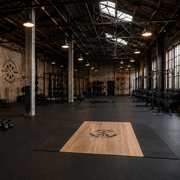 Industrial-style local gym and fitness center featuring weightlifting racks, kettlebells, medicine balls, and a wooden lifting platform in a spacious warehouse facility with brick walls and natural lighting.
