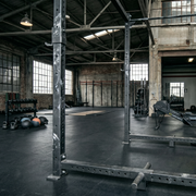 An industrial-style gym with a spacious, open floor plan and high ceilings. The room features dark rubber flooring, exposed brick and concrete walls, and large multi-paned windows that let in natural light. Multiple black power racks are positioned in the foreground and middle ground. Various pieces of workout equipment are scattered throughout the space, including rowers, kettlebells, medicine balls, and a pull-up bar rig in the background. 