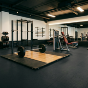 An interior view of a modern, spacious gym featuring dark rubber flooring and white walls. The room is equipped with various strength training gear.