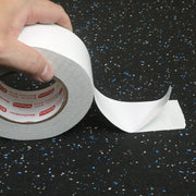 Hand holding a roll of double sided tape with a strip sticking to a blue speckled rubber flooring. 