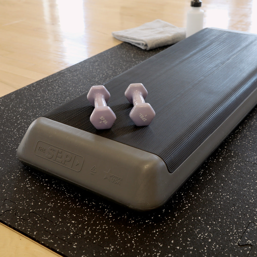 Step platform with dumbbells on top of 6 interlocked rubber gym tiles forming a mat. The rubber tiles are lying of top of a wood floor.