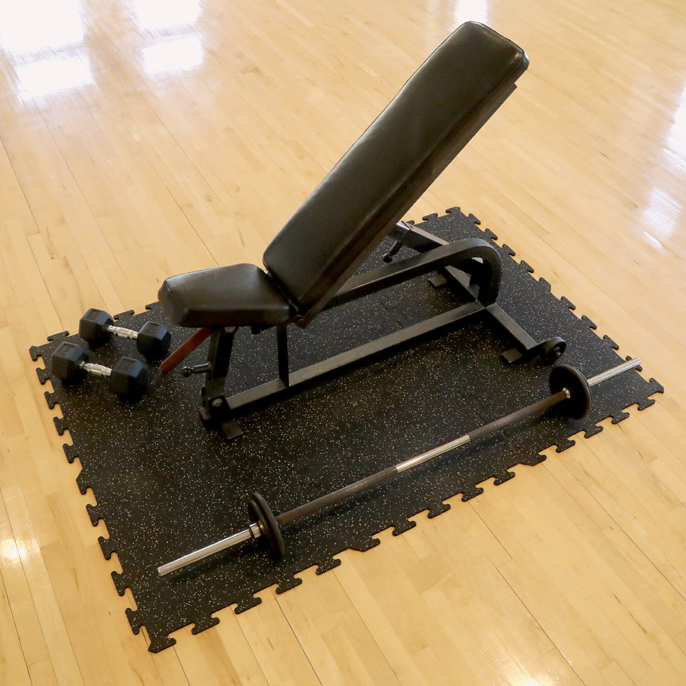 Gym bench, barbell, and dumbbells on top of two interlocked rubber gym tiles over a wooden floor.